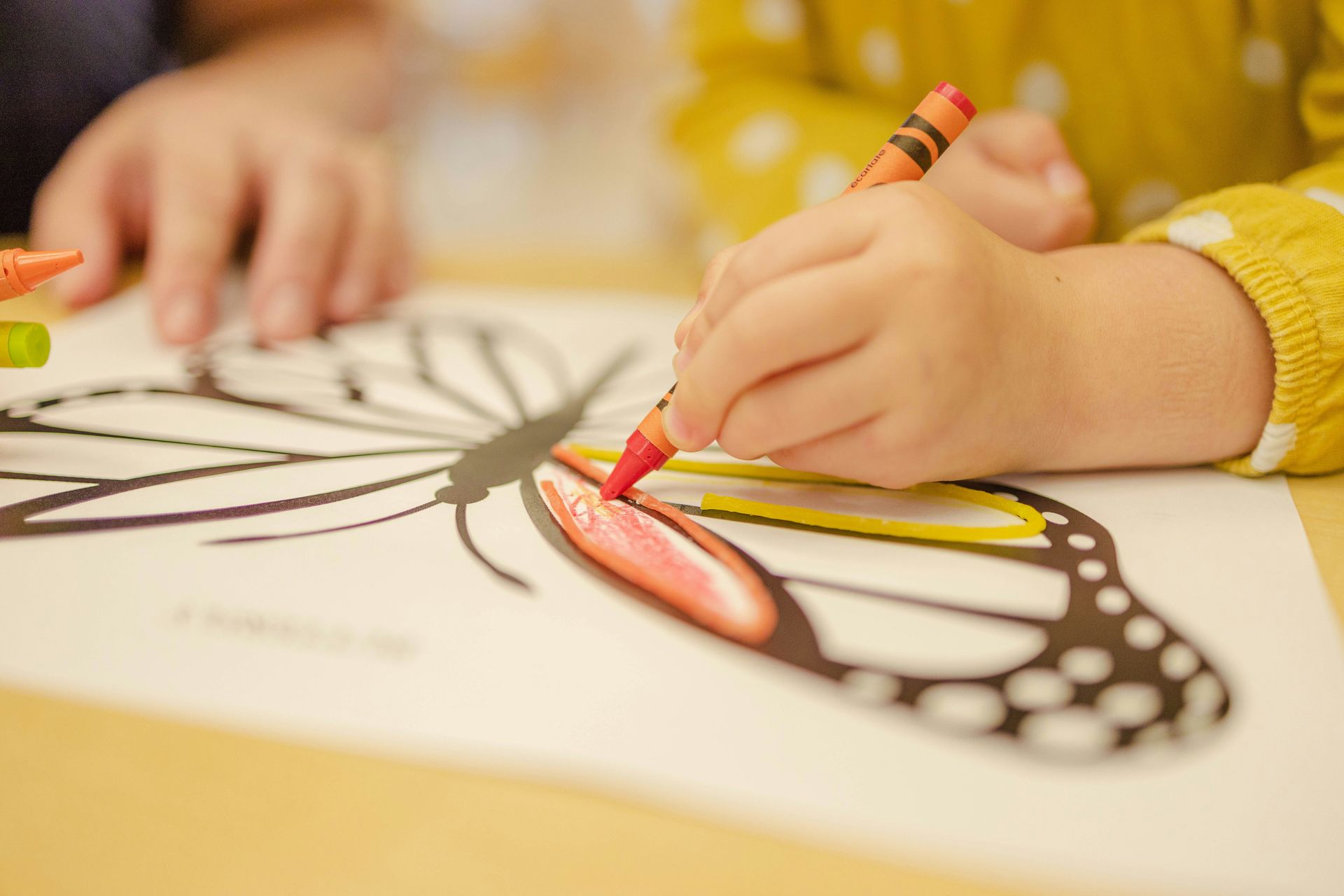A child coloring a butterfly drawing with a red crayon on white paper